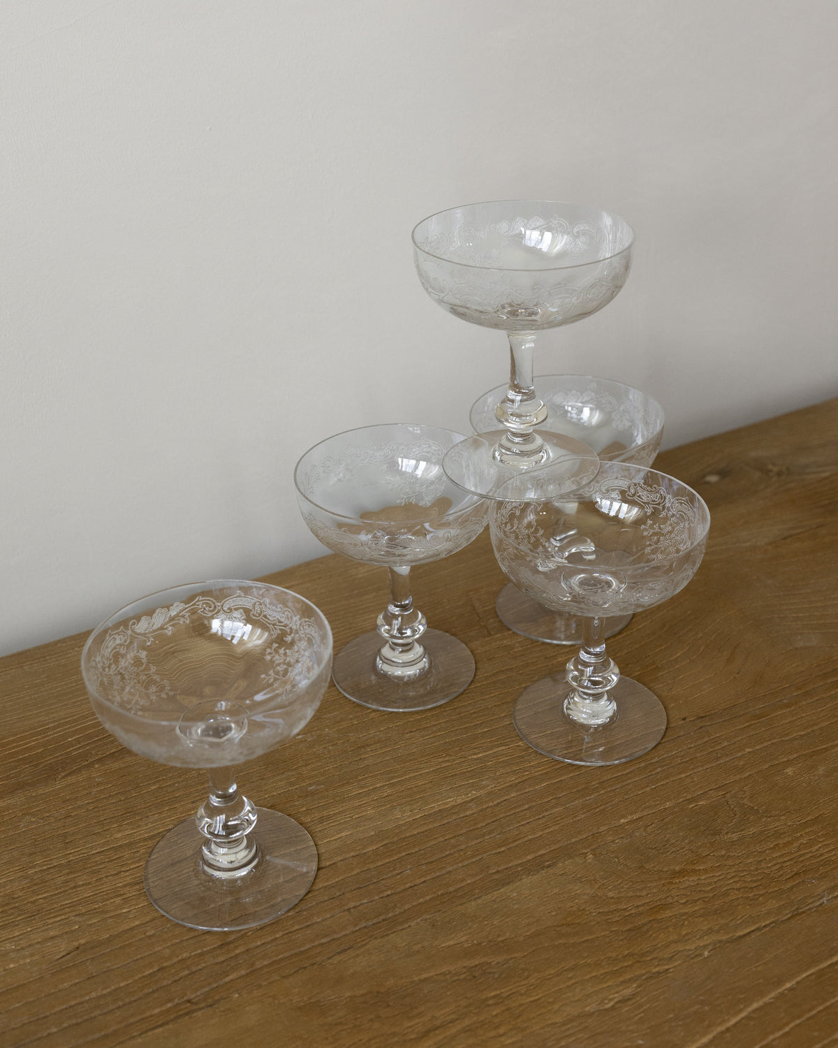 Four clear cut-crystal champagne coupe glasses arranged on a wooden tabletop against a plain light wall.