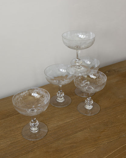 Four clear cut-crystal champagne coupe glasses arranged on a wooden tabletop against a plain light wall.