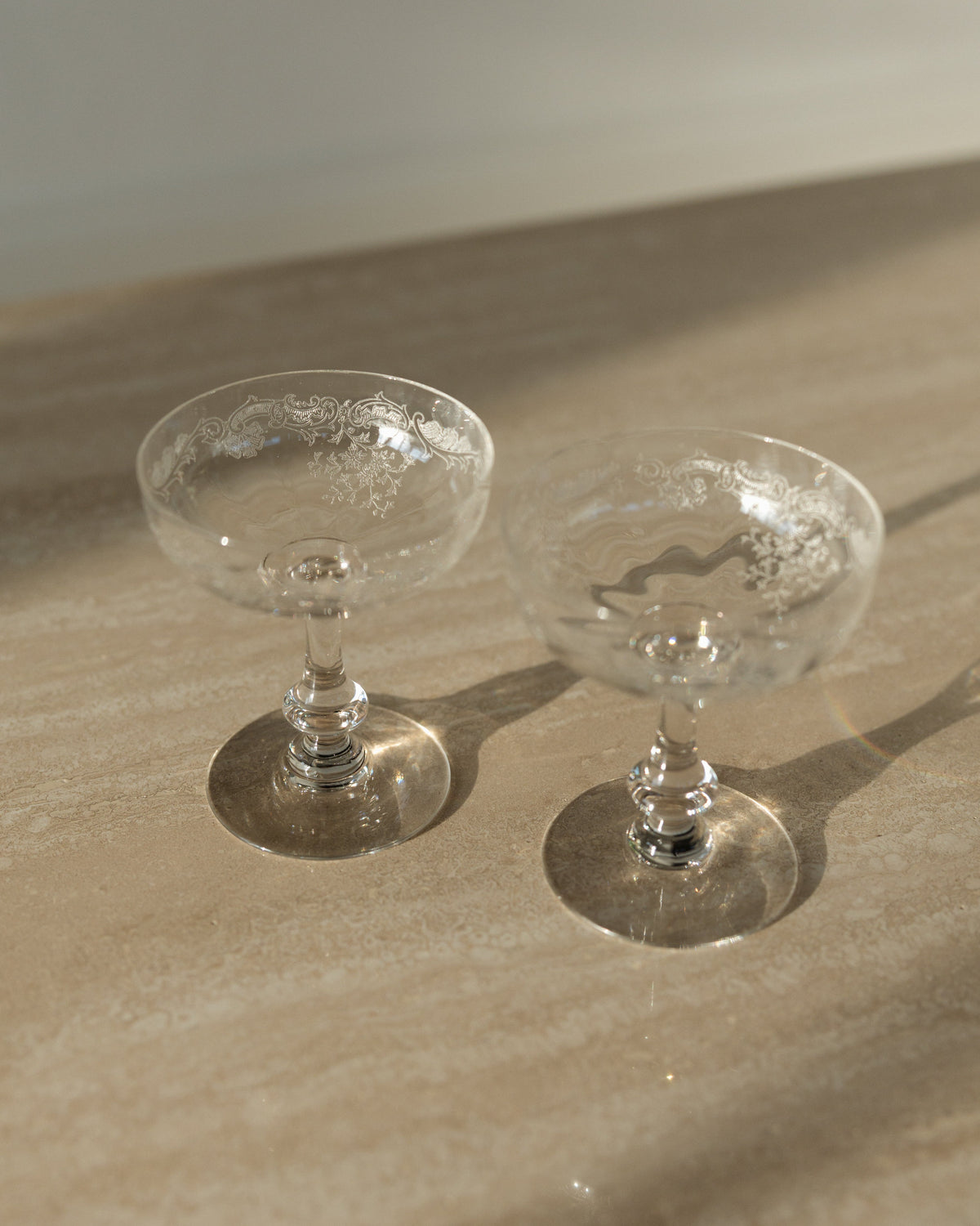 Two clear glass coupe cups with textured interiors, resting on a beige stone surface under warm natural sunlight.