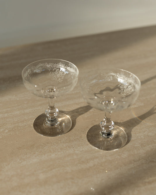 Two clear glass coupe cups with textured interiors, resting on a beige stone surface under warm natural sunlight.