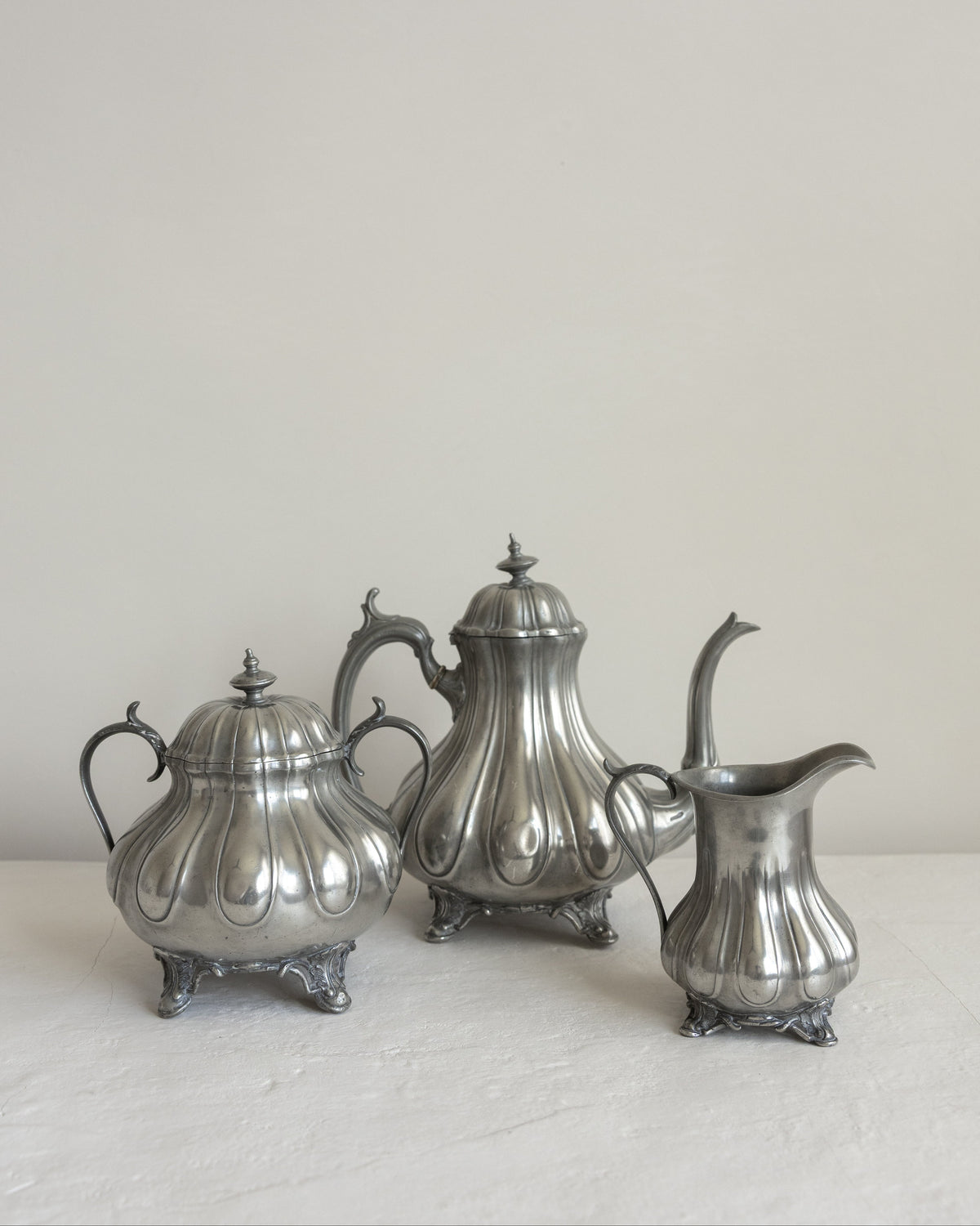 Three vintage silver metal tea and coffee serving pieces, including a teapot, sugar bowl, and creamer, arranged on a light surface against a neutral off-white background.