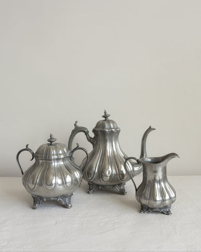 Three vintage silver metal tea and coffee serving pieces, including a teapot, sugar bowl, and creamer, arranged on a light surface against a neutral off-white background.