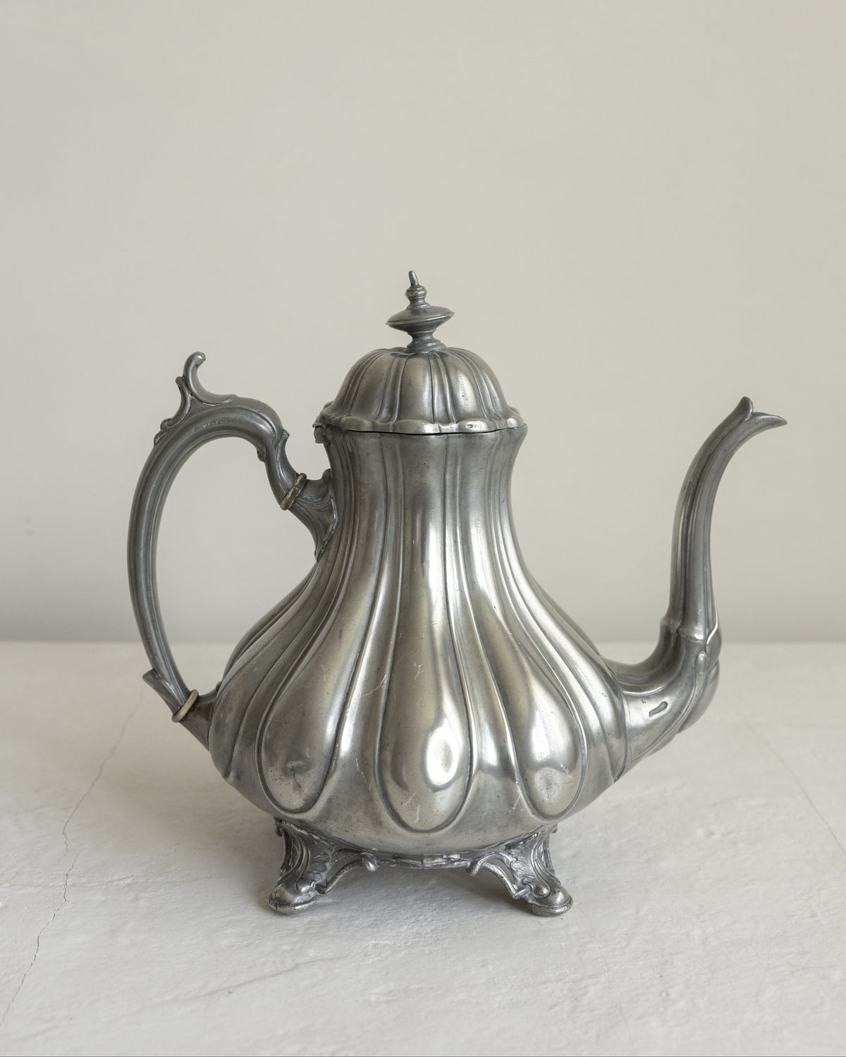 An ornate silver metal teapot with a ribbed, tulip-like body, decorative handle, and curved spout, photographed against a plain light background.
