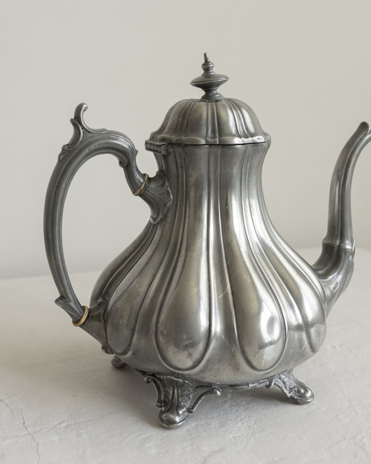 An antique silver teapot with a fluted, ribbed body, ornate handle, and small feet, shown on a neutral light gray background.