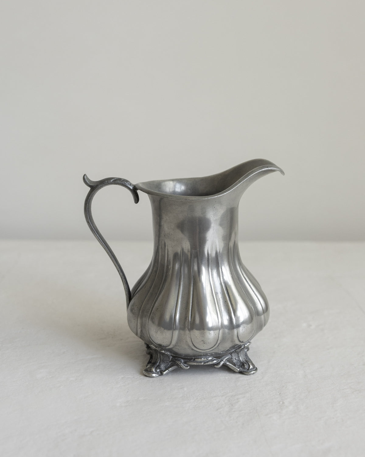 A vintage silver pewter pitcher with a fluted body, curved handle, and small decorative feet, set against a neutral light gray background.