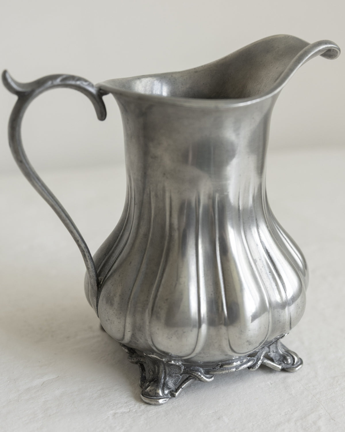 A vintage silver pewter pitcher with a fluted body, curved handle, and decorative footed base, photographed on a light neutral surface.