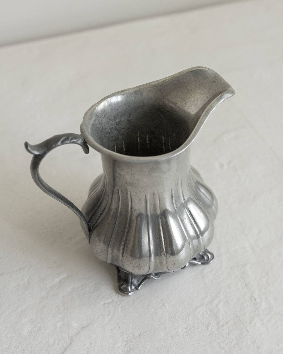 An antique silver pewter creamer pitcher with a fluted body, decorative handle, and small feet, shown on a textured off-white surface.