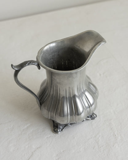An antique silver pewter creamer pitcher with a fluted body, decorative handle, and small feet, shown on a textured off-white surface.