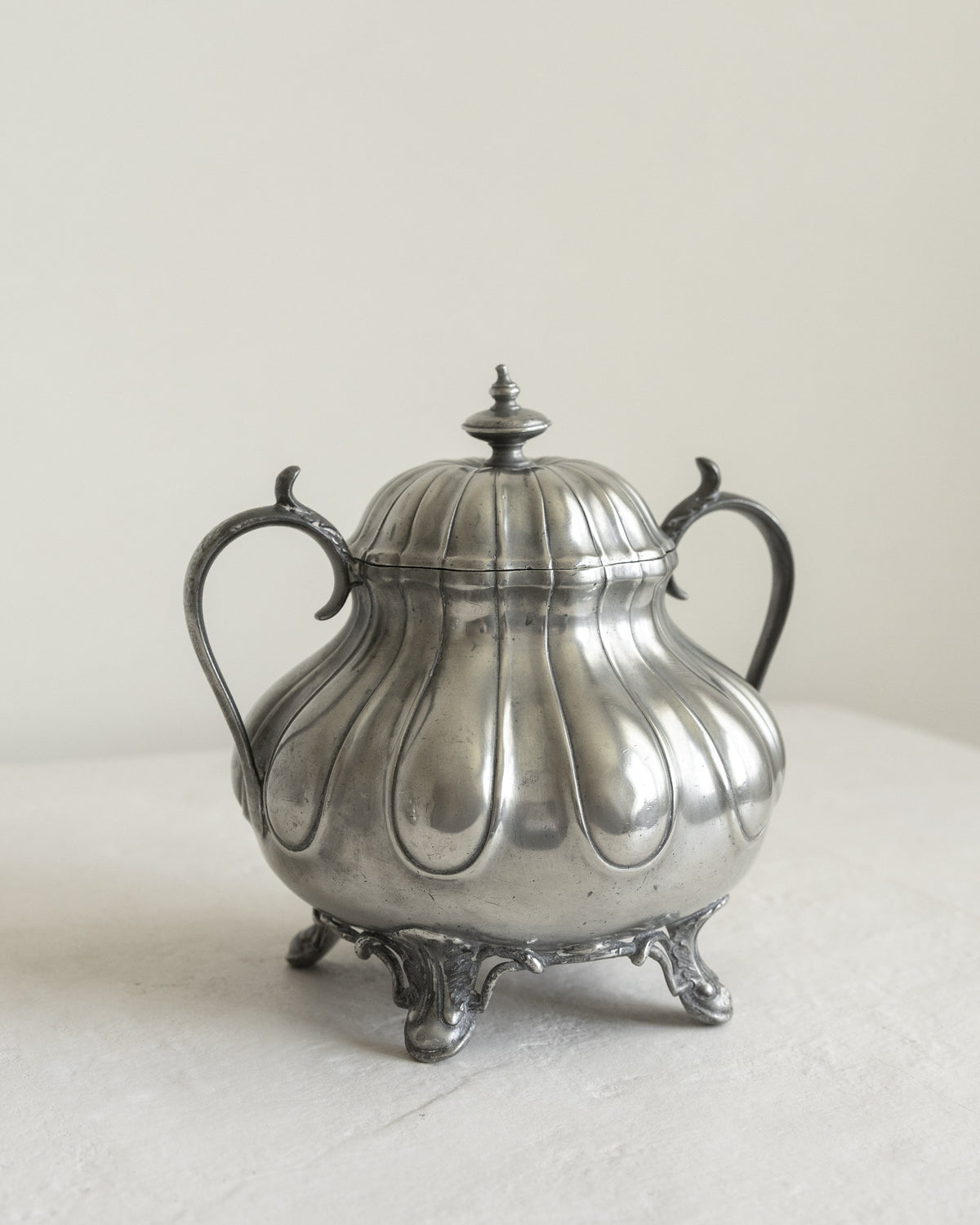 A vintage silver-toned metal lidded sugar bowl or caddy with ornate handles and feet, photographed on a light neutral surface against a plain off-white background.