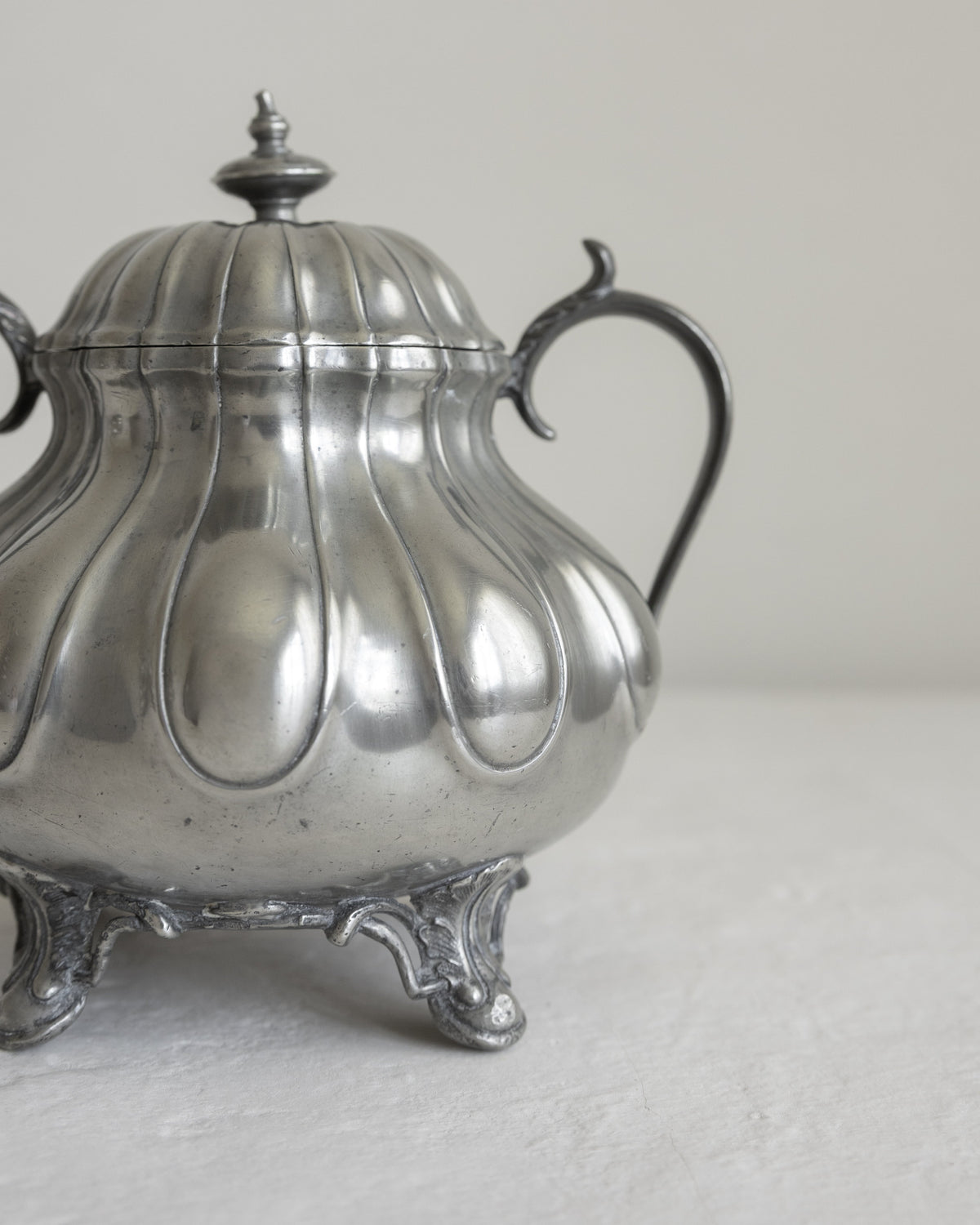 A vintage silver-toned metal teapot or sugar bowl with a ribbed lid, curved handle, and ornate feet, photographed against a plain light background.