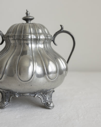 A vintage silver-toned metal teapot or sugar bowl with a ribbed lid, curved handle, and ornate feet, photographed against a plain light background.