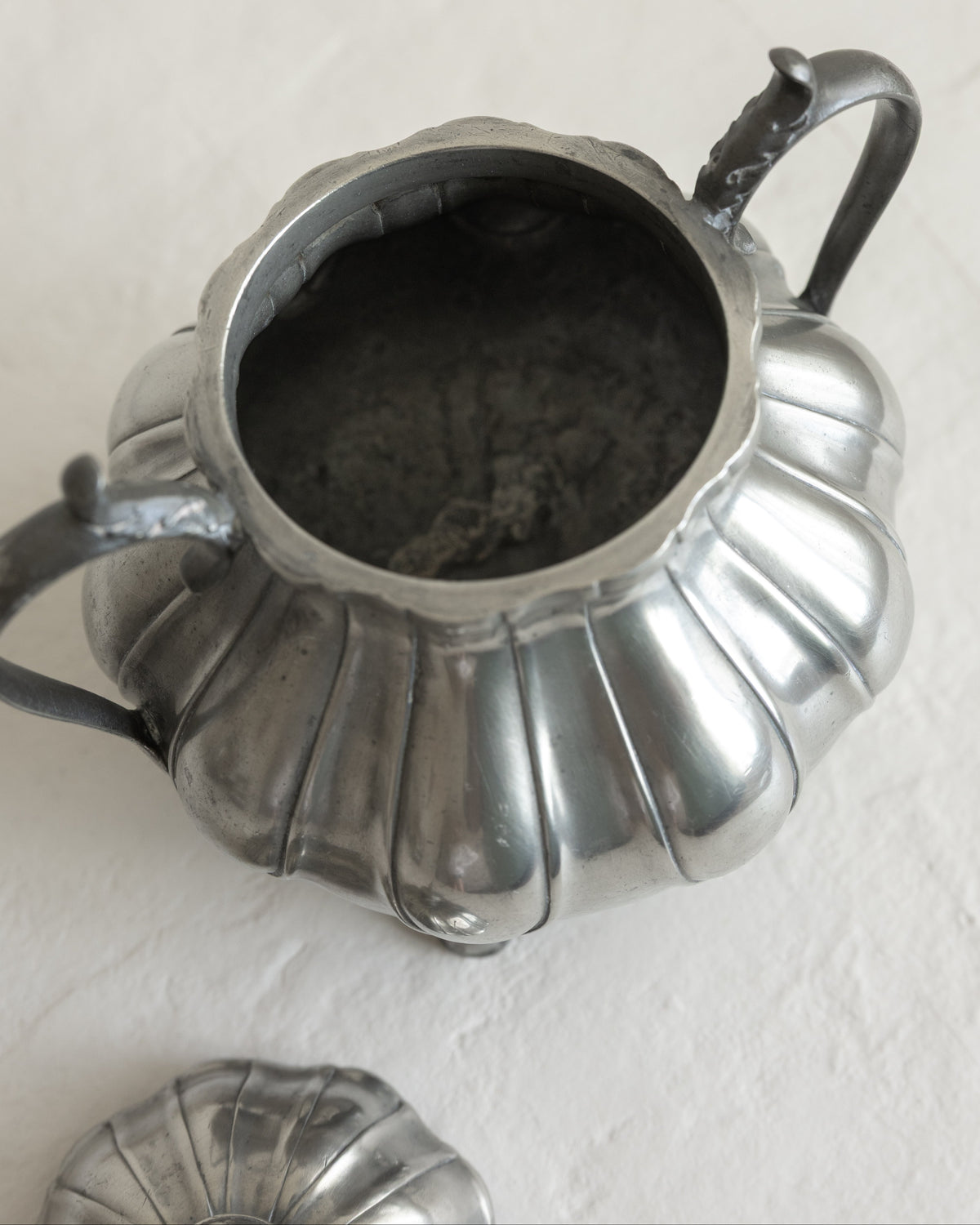 A vintage silver-toned metal teapot with a ribbed, pumpkin-like body and a matching lid partially visible at the bottom, set against a textured off-white background.