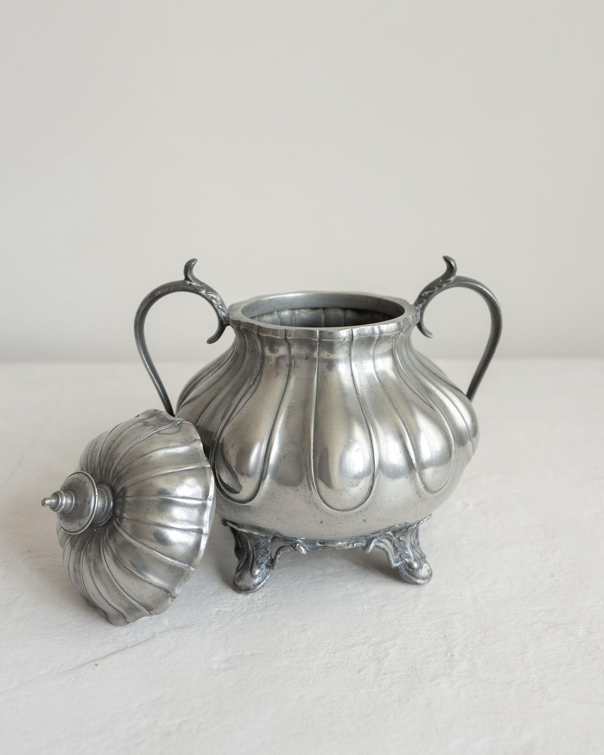 A vintage silver-toned metal sugar bowl with a matching lid, featuring a ribbed, fluted body, two curved handles, and three feet, set against a plain off-white background.