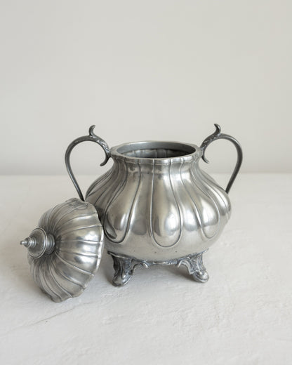 A vintage silver-toned metal sugar bowl with a matching lid, featuring a ribbed, fluted body, two curved handles, and three feet, set against a plain off-white background.