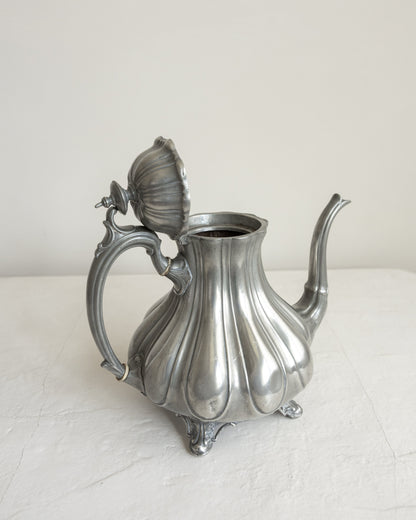 An ornate silver pewter teapot with a ribbed, pumpkin-like body, a decorative handle, and a lid shaped like a flower bud, set against a plain off-white background.