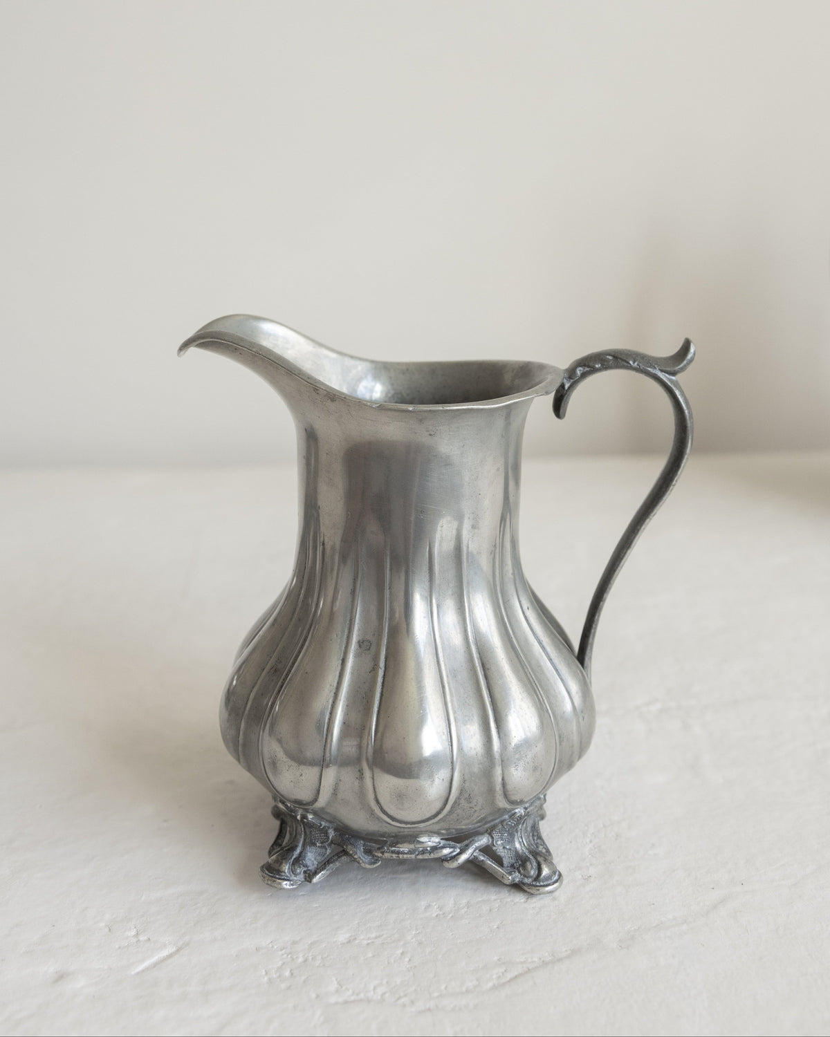 A vintage silver pewter pitcher with a fluted body, ornate footed base, and curved handle, set against a neutral off-white background.