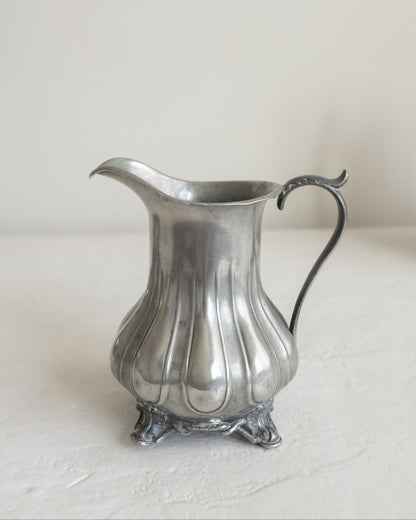 A vintage silver pewter pitcher with a fluted body, ornate footed base, and curved handle, set against a neutral off-white background.
