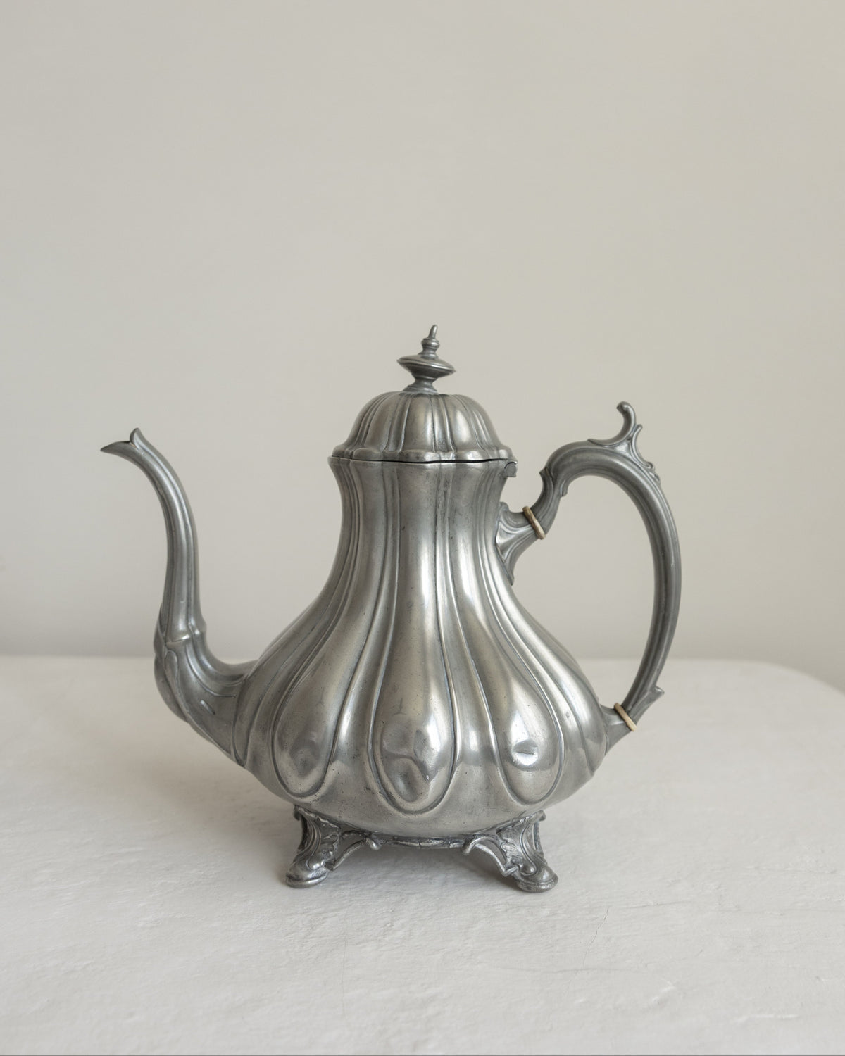 A vintage silver teapot with a fluted, lotus-like body, a curved spout, and an ornate handle, resting on a white surface against a neutral beige background.
