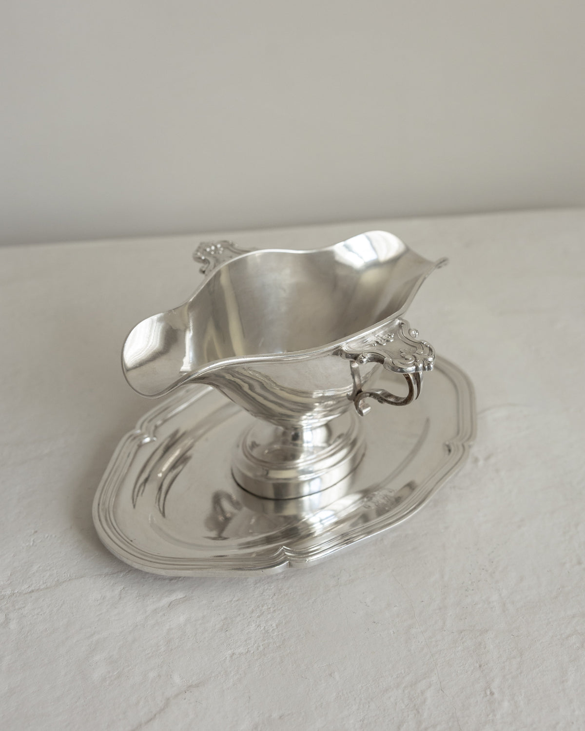 A polished silver-toned metal gravy boat with a matching saucer, photographed on a light neutral surface against a plain light gray background.