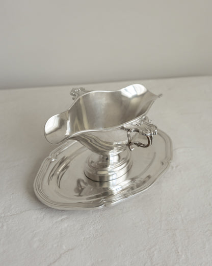 A polished silver-toned metal gravy boat with a matching saucer, photographed on a light neutral surface against a plain light gray background.