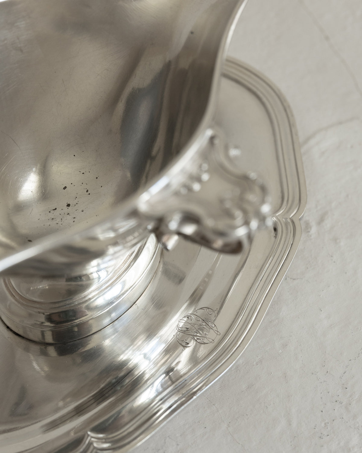A close-up of a polished silver metal pitcher resting on a matching scalloped silver saucer against a light textured background.