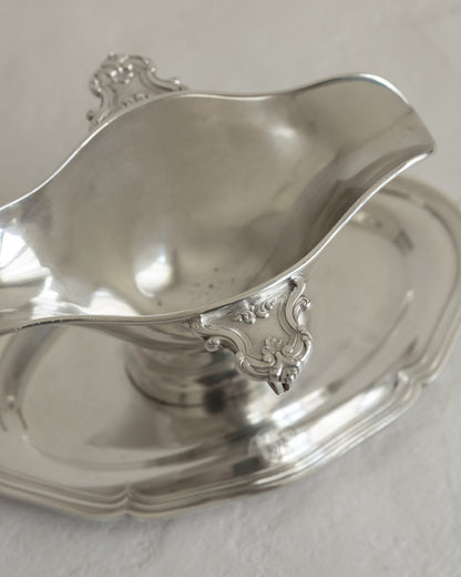 A polished silver gravy boat with ornate scrollwork handles, shown resting on a matching silver serving tray against a light neutral background.