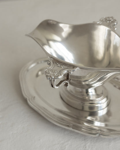 A polished silver-toned metal pitcher with ornate decorative handles, shown alongside a matching silver-toned saucer on a light neutral surface.