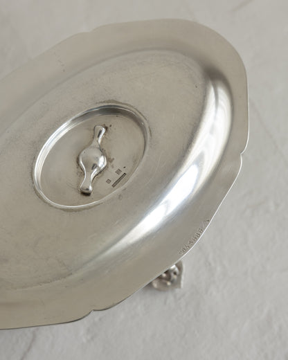 A close-up of a polished silver metal lid with a scalloped edge and a small decorative handle in the center, resting on a textured off-white surface.