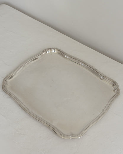 A square silver serving tray with scalloped edges, displayed on a white textured surface against a neutral light gray background.