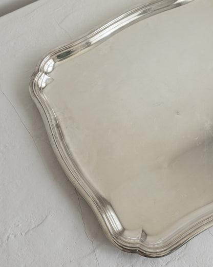 A close-up of a polished silver tray with a scalloped edge, resting on a textured off-white surface.