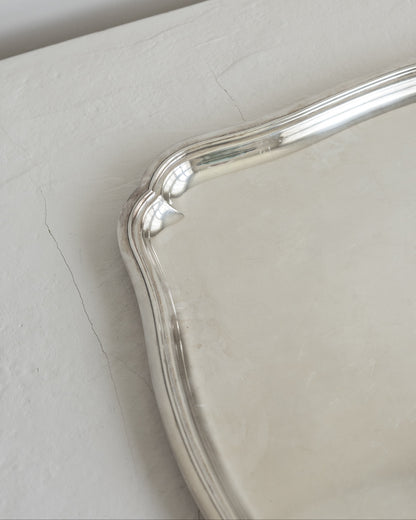 A close-up of a polished silver tray corner with a curved, decorative edge, resting on a textured off-white surface.
