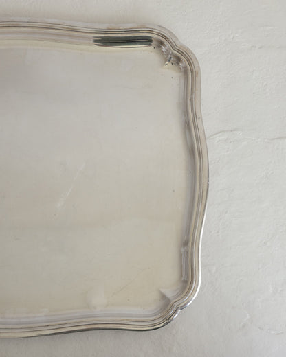 A close-up of a silver-toned metal tray with a scalloped edge, resting on a textured off-white surface.